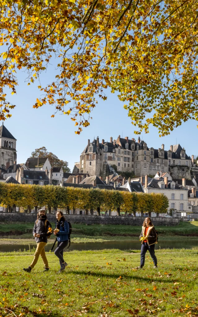Trois personnes marchent dans un parc avec des arbres à feuilles jaunes, la cité médiévale de Saint-Aignan en arrière-plan, et un ciel bleu clair.