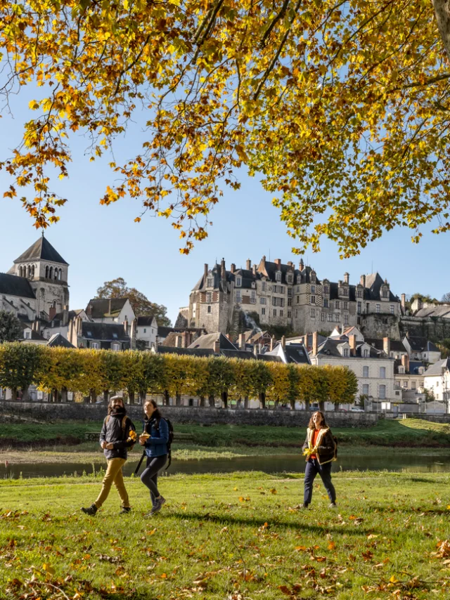 Trois personnes marchent dans un parc avec des arbres à feuilles jaunes, la cité médiévale de Saint-Aignan en arrière-plan, et un ciel bleu clair.