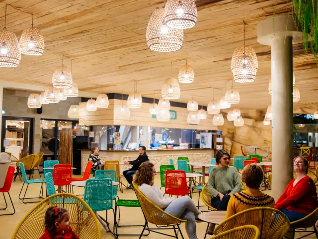 Des visiteurs assis dans un espace détente aux couleurs vives, avec des chaises en rotin, un plafond en bois et des luminaires suspendus.