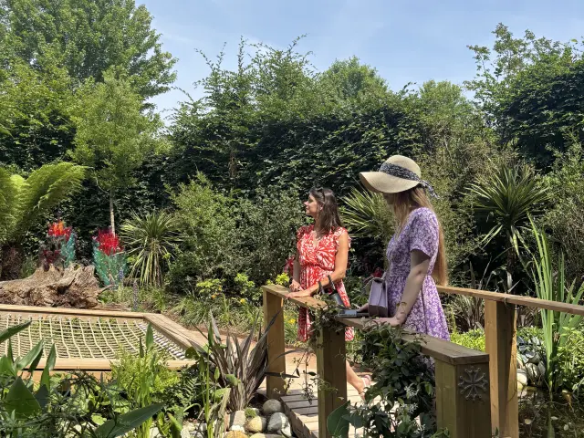 Deux femmes se tiennent sur un petit pont en bois dans un jardin luxuriant, entourées de plantes vertes et de fleurs sous un ciel bleu clair.