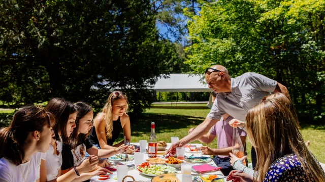 L'image capture un moment convivial d'un déjeuner en plein air, où un groupe de personnes partage un repas autour d'une table bien garnie dans un cadre verdoyant et ensoleillé.