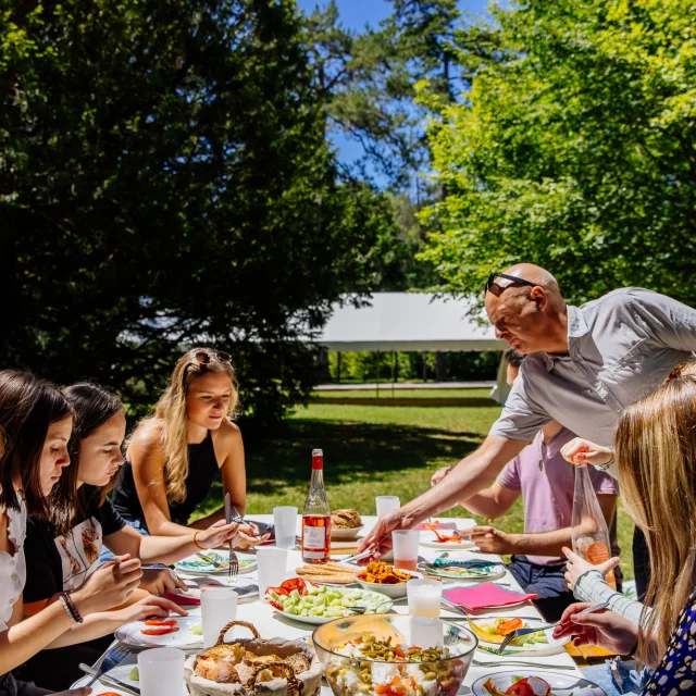 L'image capture un moment convivial d'un déjeuner en plein air, où un groupe de personnes partage un repas autour d'une table bien garnie dans un cadre verdoyant et ensoleillé.