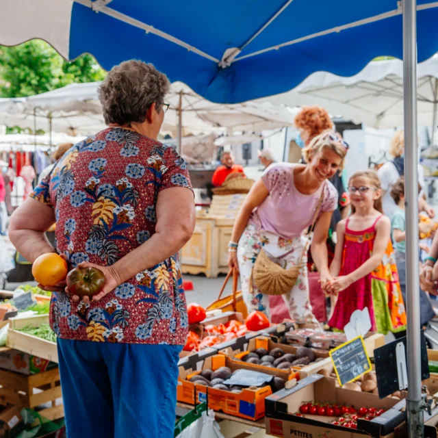 Une scène vivante dans un marché en plein air où une femme de dos tient deux tomates anciennes pendant qu'une autre femme et des enfants sourient devant un étal rempli de légumes frais.
